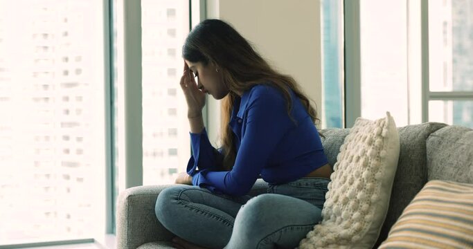Depressed lonely young 20s Indian girl sitting on home sofa, looking at window with sad thoughts, touching head, forehead, thinking over troubles, loss, facing depression, stress, emotional crisis