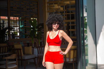 Slender middle-aged woman with curly black hair red summer clothes wearing sunglasses.