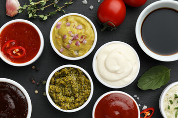 Different tasty sauces in bowls and ingredients on black table, flat lay