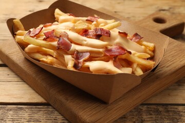 Tasty potato fries, cheese sauce and bacon in paper container on wooden table, closeup