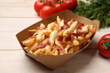 Tasty potato fries, cheese sauce, bacon and products in paper container on light wooden table, closeup