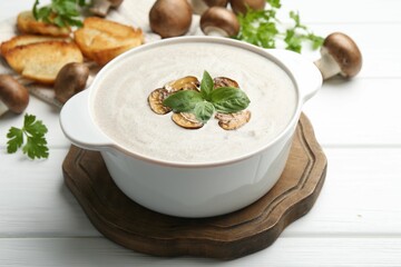 Delicious homemade mushroom soup in ceramic pot, croutons, fresh champignons and parsley on white wooden table
