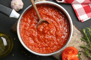Homemade tomato sauce in pot, spoon and fresh ingredients on dark table, flat lay