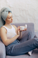 Young woman hold creme jar relaxing on couch, head wrapped in towel