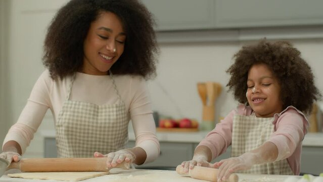 Cheerful African American mother woman teach little daughter child kid girl make flattening dough with rolling-pins in kitchen happy family use ecological products ingredients cooking together smiling