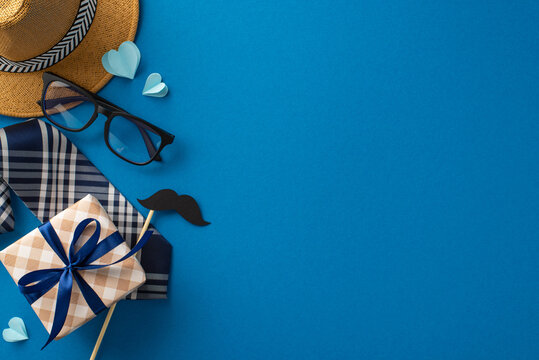 Show Dad your love and appreciation with tasteful top view setup featuring straw hat, stylish necktie, adorned gift box, glasses, mustache, and paper hearts on blue backdrop, perfect for greeting text