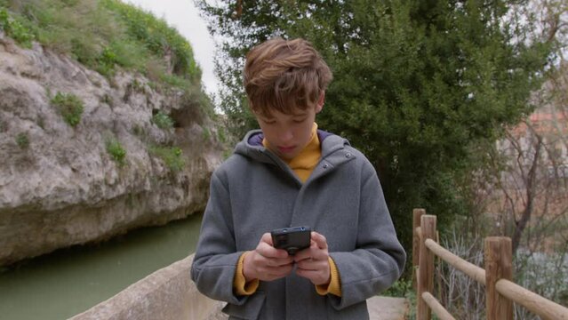 Teenager strolls by a river's edge against a backdrop of mountains, absorbed in using his phone