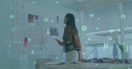Image of connected icons and numbers over biracial woman examining design of dress in office