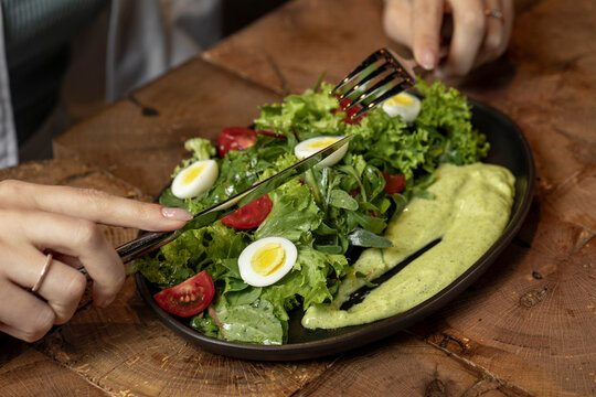 Person eating Fresh green salad with quail eggs, cherry tomatoes and guacamole in black plate on grey background