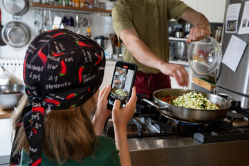 Girl filming the father who is cooking with her mobile phone. Little girl dressed in apron and...