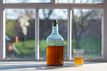 Big bottle with a drink made from fermented birch sap on the windowsill on a warm spring day, closeup. Traditional Ukrainian cold barley drink kvass in a glass jar and glass on table near yard - obrazy, fototapety, plakaty