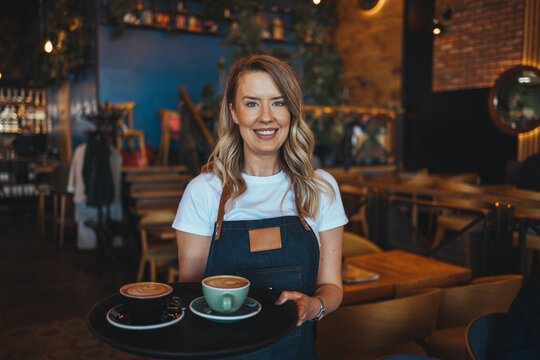 Cheerful young waitress wearing apron laughing looking at camera, happy businesswoman small business owner of girl entrepreneur cafe employee posing in restaurant coffee shop interior, portrait