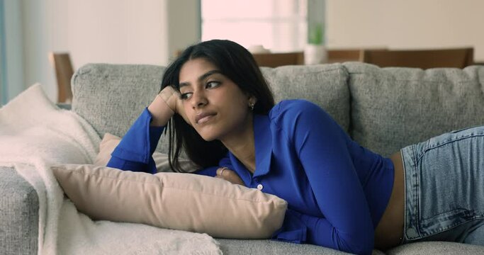 Tired sleepy young Indian woman falling asleep on soft comfortable home couch, lying on belly, looking away, closing eyes. Sad calm attractive 20s girl thinking on bad news, troubles