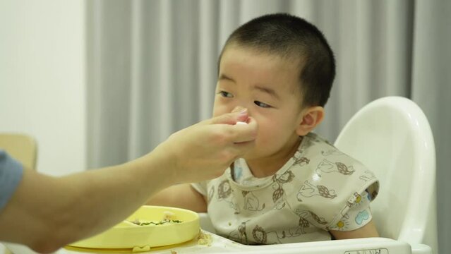 Close of mother hand feeding Asian baby boy, sitting on white highchair at home