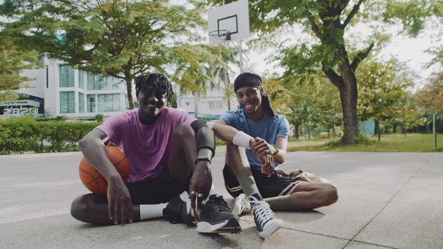 Portrait of male basketball players in activewear sitting on outdoor city court and smiling at camera
