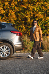a smiling woman walks next to her car on the background of an autumn forest