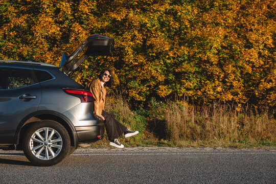a smiling woman sits in the trunk of a car on the background of an autumn forest