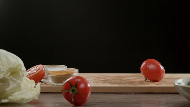 Few juicy and ripe red tomatoes drop onto the chopping board, among other vegetables that are lying on the table. Splashes of water fly around as they land. Close up. Slow motion.