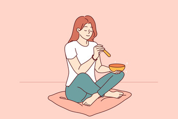Woman practices tibetan meditation using golden singing bowl, sits on mat in room with smile on face. Girl enjoys buddhist meditation that cleanses chakra and improves mood or harmonizes soul - 785545977