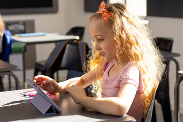 In school, in classroom, young Caucasian girl with curly blonde hair is using a tablet - 785549322