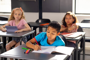 In school, in classroom, boy in blue shirt writing in notebook, two girls watching - 785549325