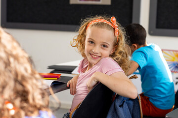 In school, young Caucasian girl with curly hair is smiling, sitting at a desk in a classroom