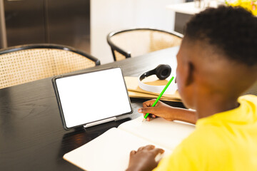 African American boy writing in notebook at home, tablet with blank screen nearby with copy space - 785549542