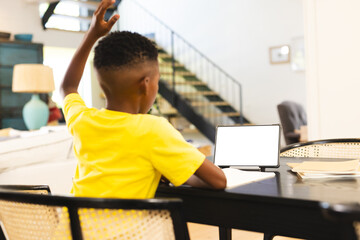 African American boy sitting at table, raising hand near tablet at home with copy space - 785549761