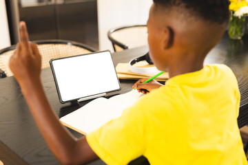 African American boy focusing on writing at home with copy space, tablet with blank screen nearby
