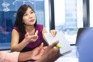 Asian woman discussing with colleague, both using laptops in a modern business office - 785549773