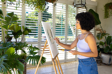 Young biracial woman painting on canvas in plant-filled room at home