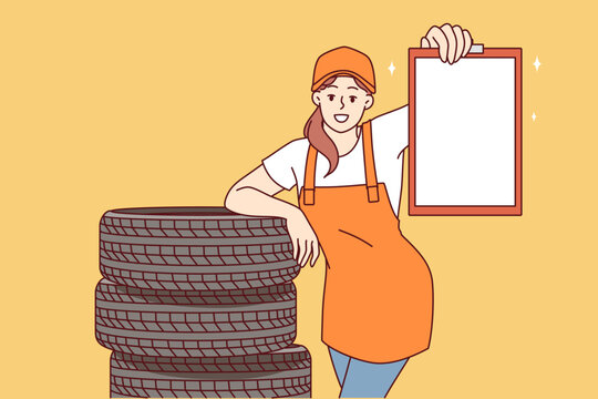 Woman mechanic from car repair shop stands near replacement tires for automobile wheels and demonstrates empty clipboard. Copy space in hands of girl mechanic in overalls for repairing vehicles