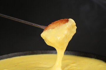 Dipping piece of bread into fondue pot with tasty melted cheese against dark gray background,...