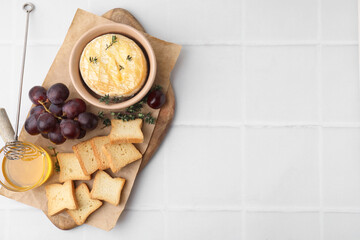 Tasty baked camembert in bowl, croutons, grapes, honey and thyme on white tiled table, top view. Space for text
