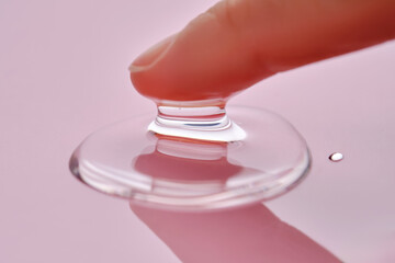 A finger touches a drop of cosmetic product on a pink background.