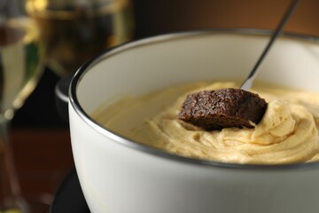 Dipping piece of bread into fondue pot with melted cheese on dark background, closeup