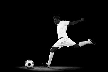 Creative image of young man in white uniform, football player in motion with ball on black...