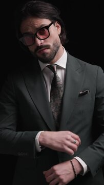 cool bearded businessman with sunglasses looking to side, adjusting suit, crossing arms and being cool with hands in pockets on black background