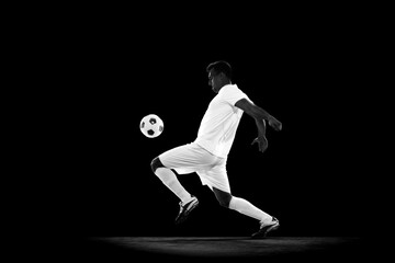 Dynamic creative image of young man, football player in white uniform in motion with ball, training on black background. Monochrome. Professional sport, game, competition, tournament, action concept