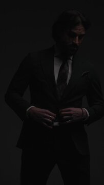 vertical video of cool fashion man adjusting elegant grey suit while looking to side in front of grey background