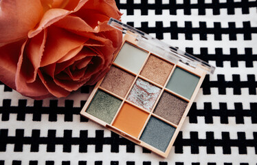 Multicolored green brown blue eye shadows palette on black and white table next to orange rosebud. Cosmetic woman's table top view. Make up product.