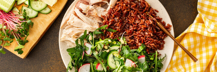 Delicious healthy breakfasts, chicken breast with red rice and salad mix with fresh seasonal organic vegetables and microgreens, top view of a plate with food and a cutting board with vegetables, bann