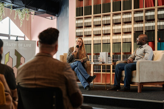 Happy young woman with open book in hands sitting in armchair and talking in microphone to public in library