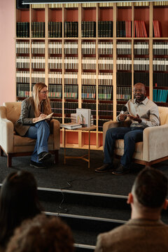 Young male novelist speaking in microphone while sitting in armchair on talk show stage against shelves with books