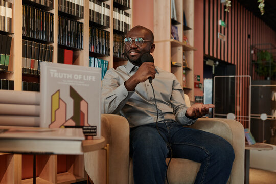 Young confident author with microphone answering question of journalist or show host while sitting in armchair