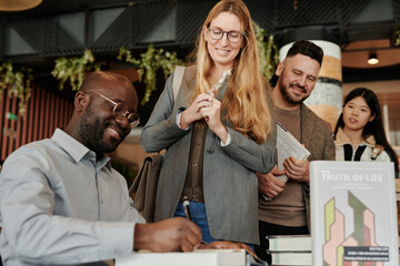 Young smiling male author signing one of books sold to fans standing in queue during autograph session in library