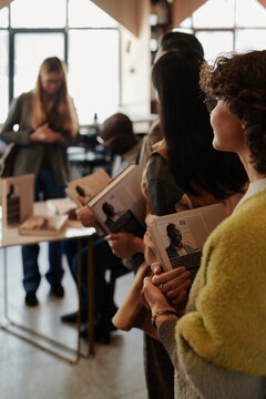Side view of young female fan with book of popular author standing in queue to get autograph after presentation