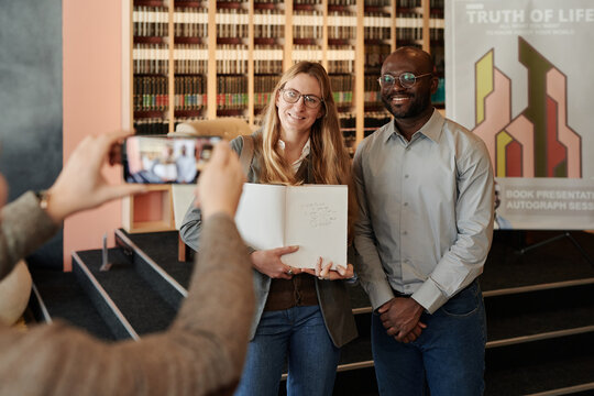 Happy young woman showing autograph of her favorite author in book while standing next to him and posing for camera