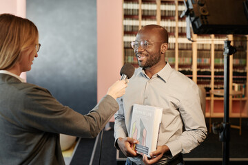 Young African American writer with his new book speaking in microphone while answering questions of journalist during interview