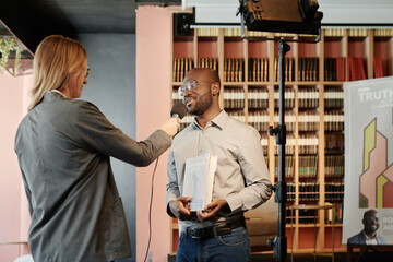 Young female journalist with microphone interviewing popular writer presenting his new book at literature event in library
